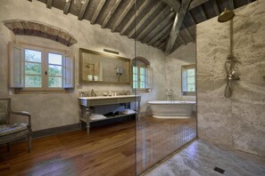 A bathroom with a wooden floor, a bathtub, a shower, a vanity with a mirror, and a chair.