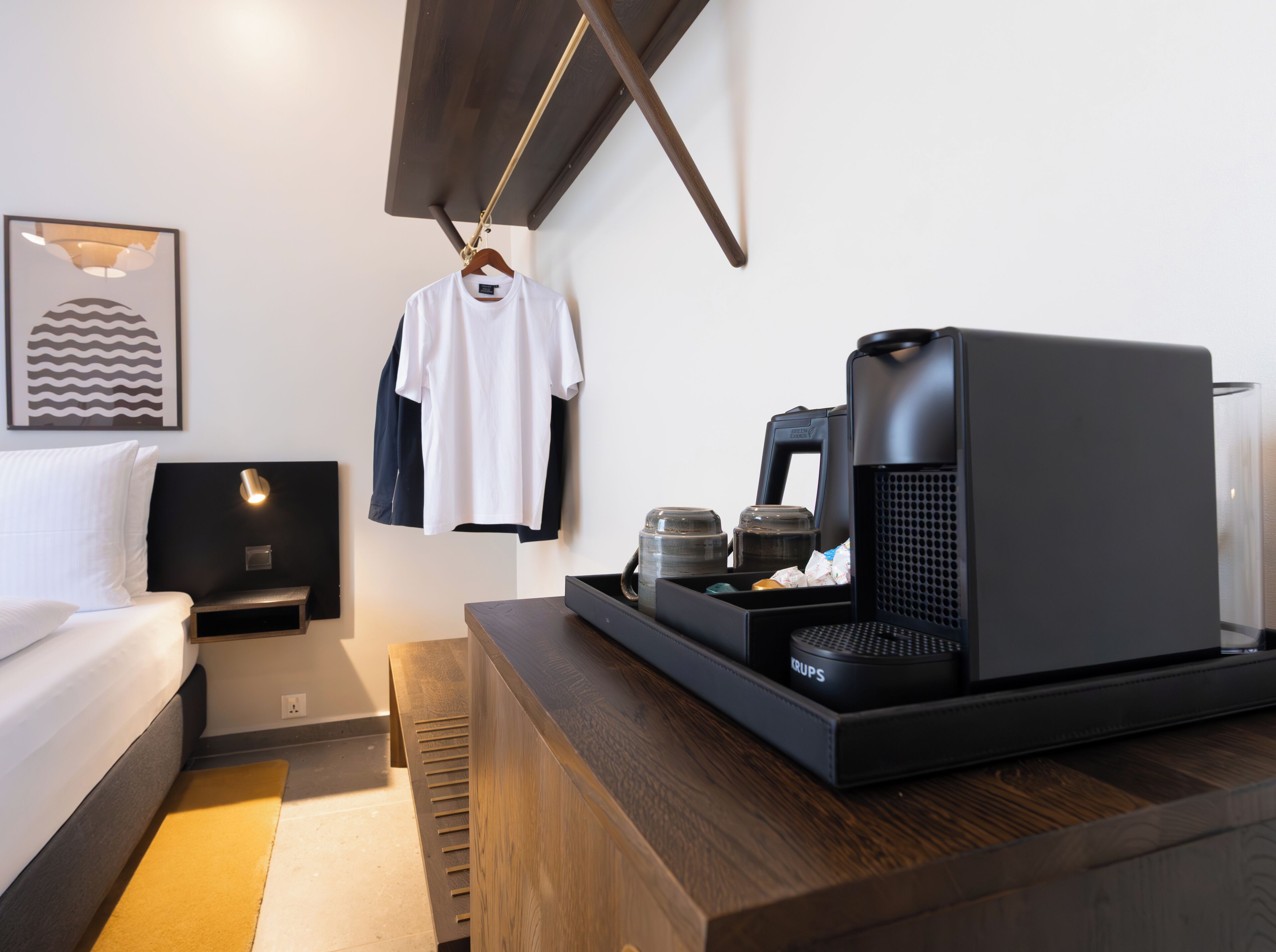 A modern hotel room with a bed, a bedside table with a coffee machine, a hanging clothes rack, and a framed artwork on the wall.