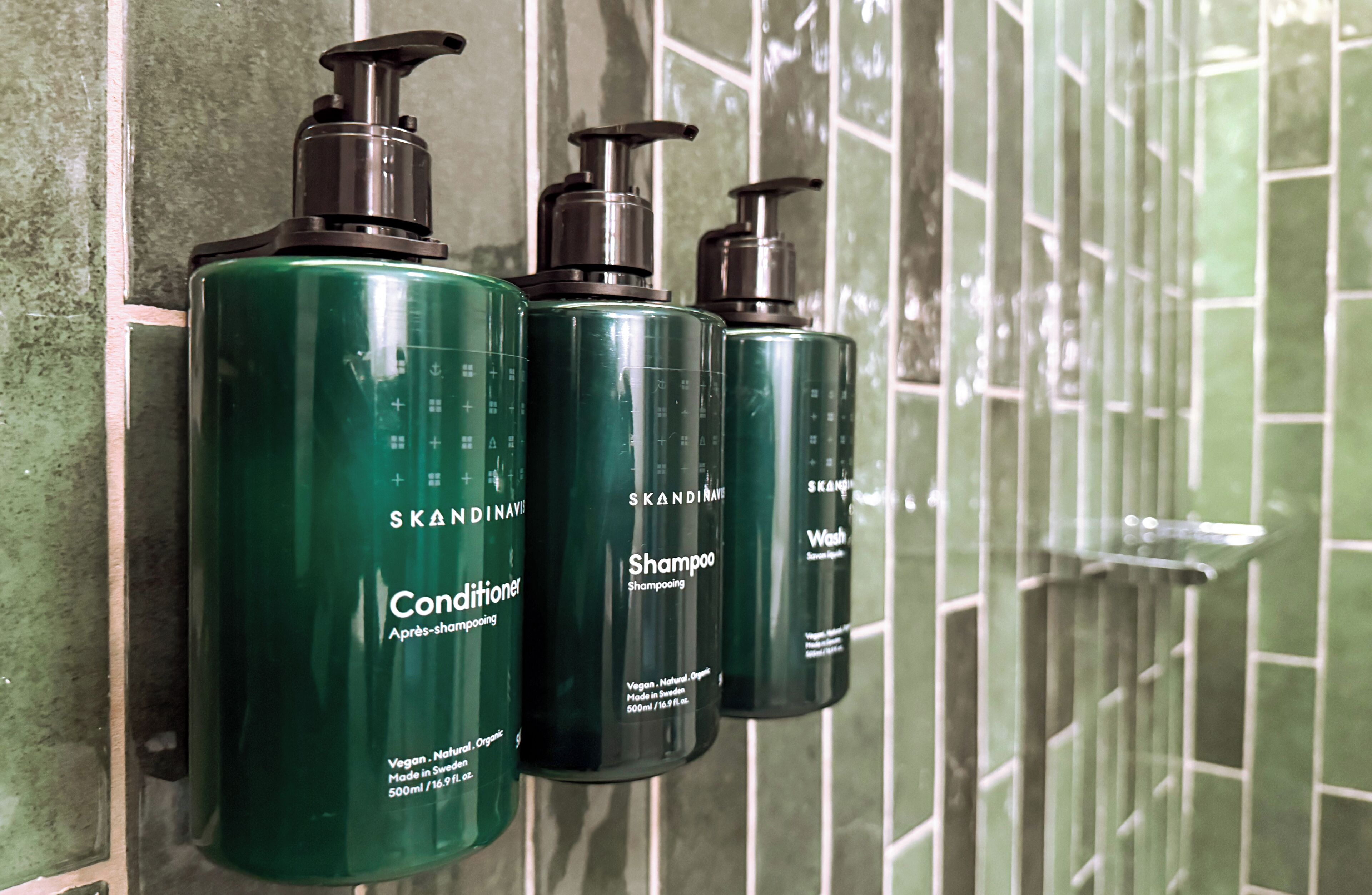 Tres botellas de champú acondicionador SKANDINAVIAN sujetas a una pared de azulejos.