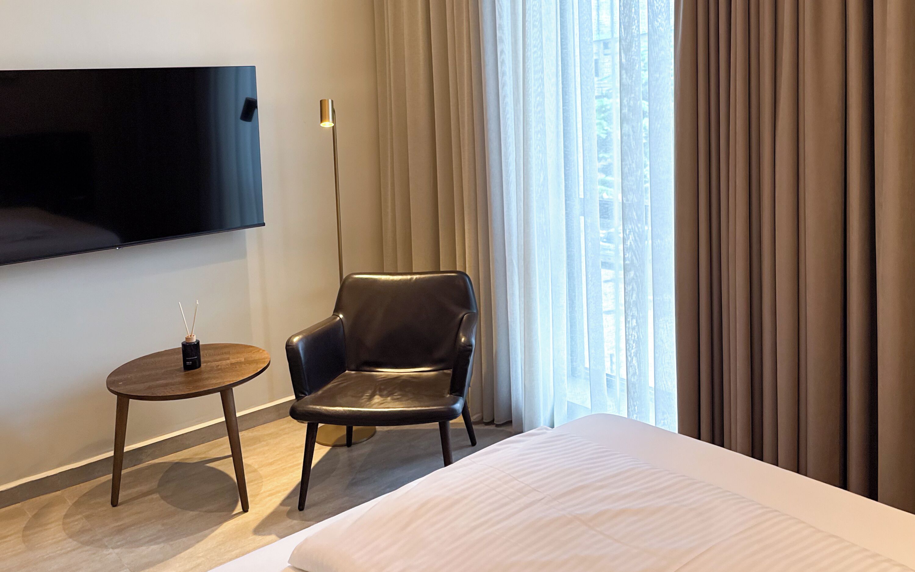 A hotel room with a bed, a leather chair, a small round table, a TV mounted on the wall, and a window with curtains.