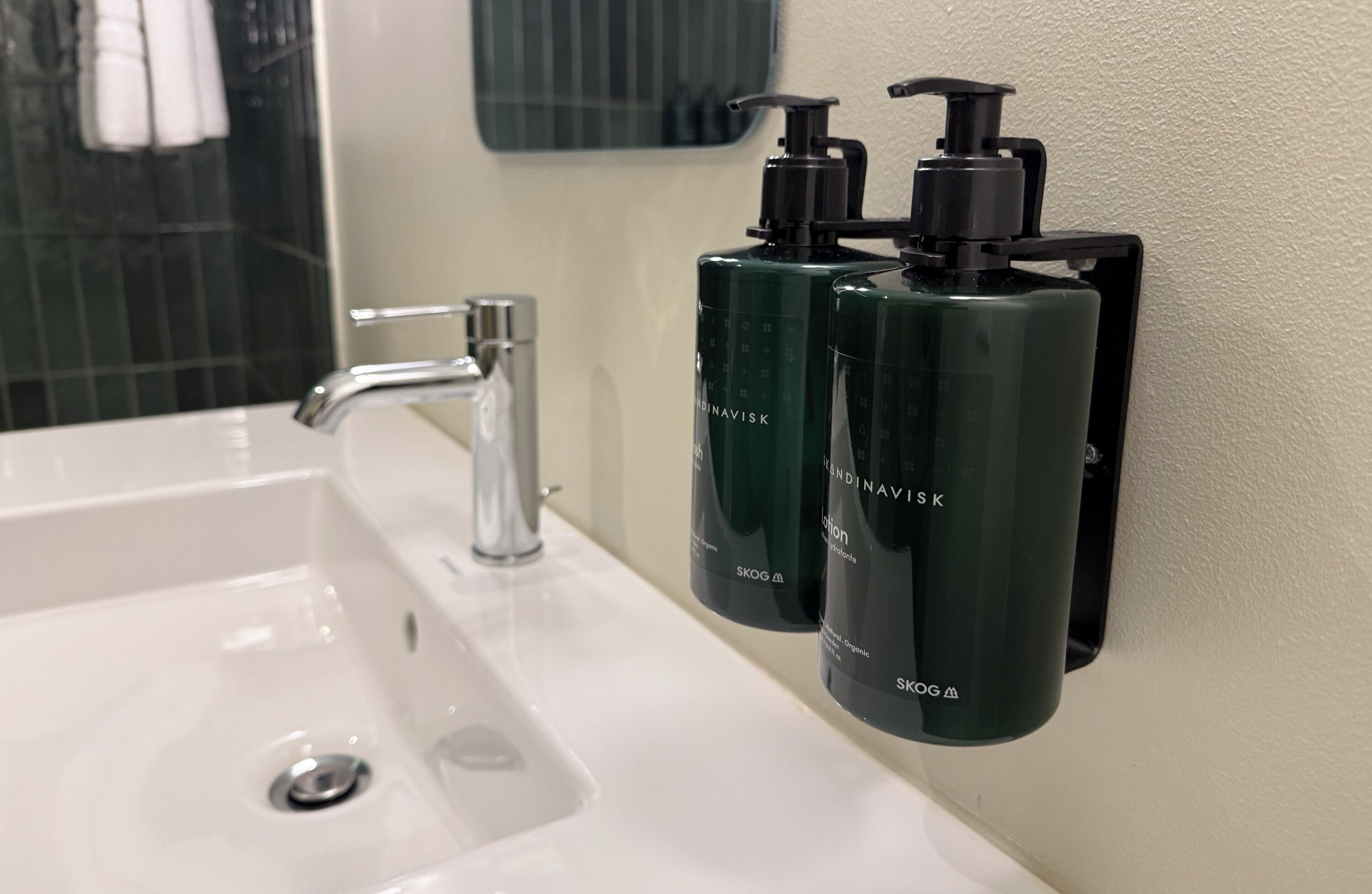 Une salle de bain avec un lavabo blanc, un robinet argenté et deux bouteilles vertes de la marque SKOG fixées au mur.
