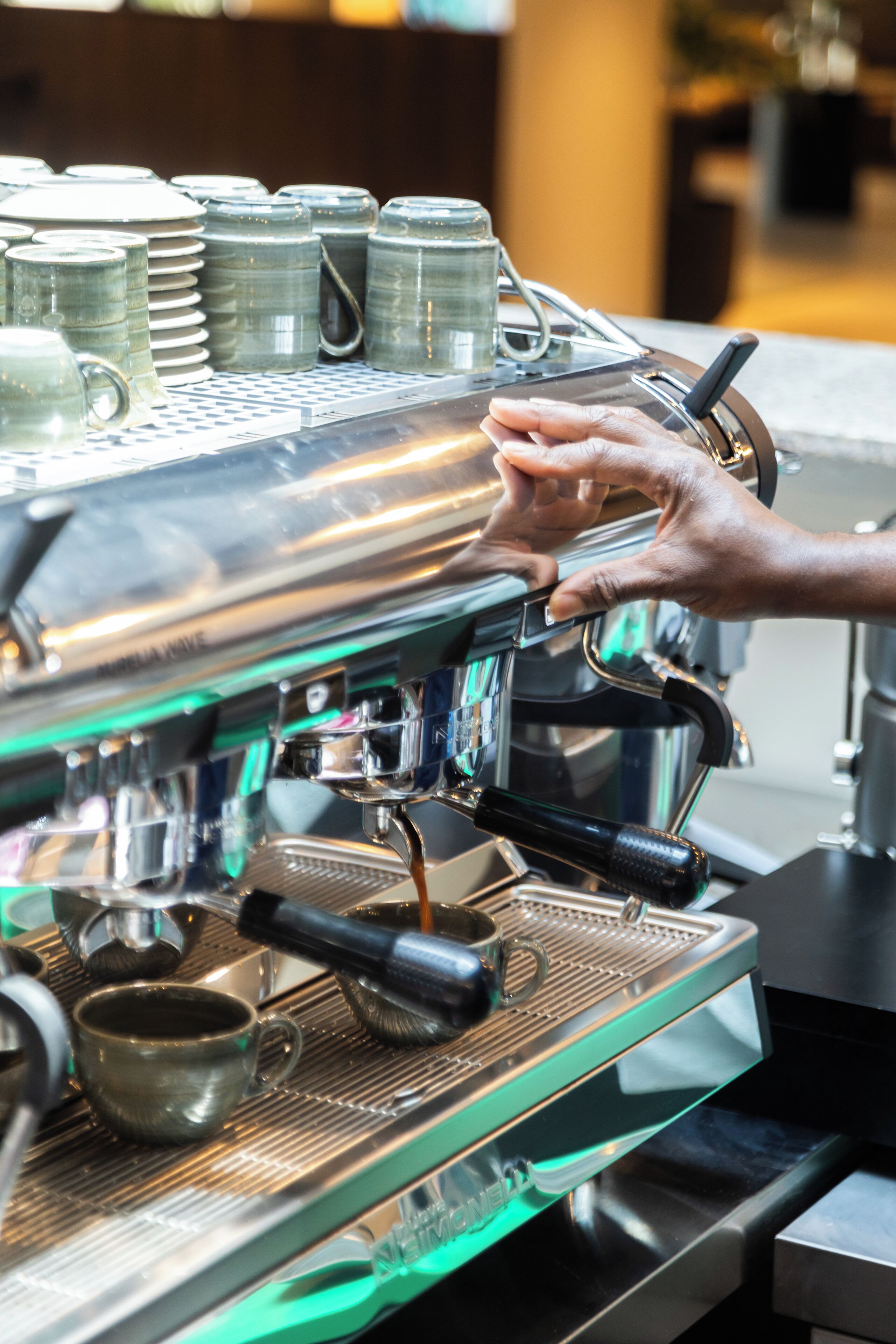 A coffee machine with a hand adjusting a lever, surrounded by stacked cups and a small cup on the counter.