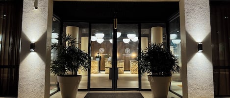 Fachada de hotel con diseño moderno, que incluye puerta de vidrio, plantas en macetas y una recepción con un logotipo.