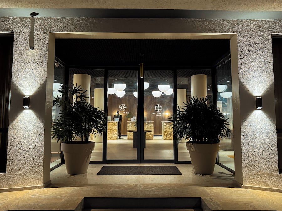 Fachada de hotel con diseño moderno, que incluye puerta de vidrio, plantas en macetas y una recepción con un logotipo.