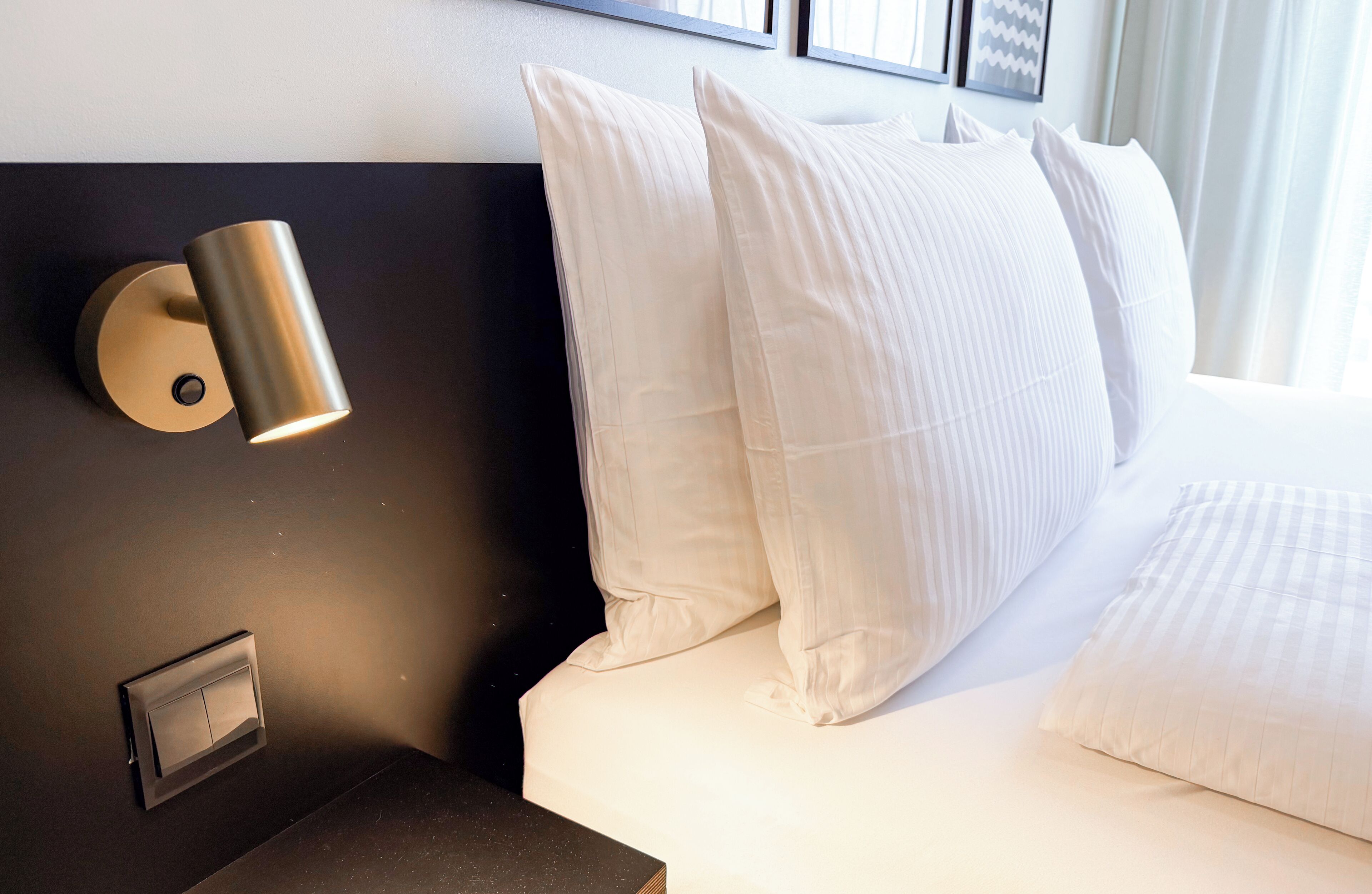 A hotel room with a modern design, featuring a bed with white linens, a dark headboard, and a wall-mounted light fixture.