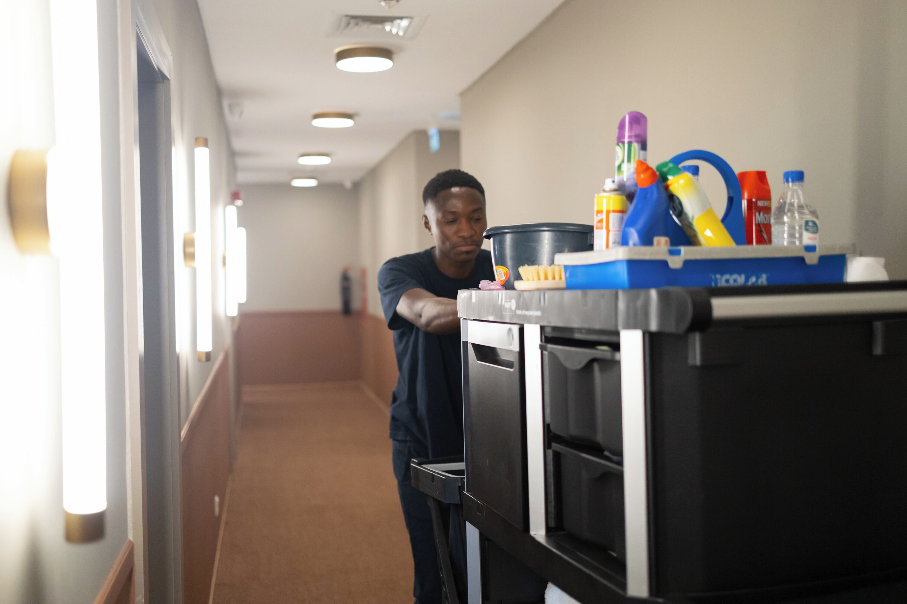 A person pushing a cart with cleaning supplies in a hallway with a carpeted floor and recessed lighting.