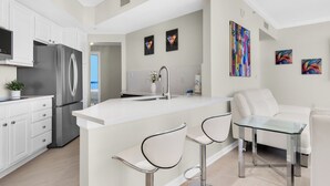 A modern kitchen with white cabinetry, a stainless steel refrigerator, a central island with bar stools, and a white sofa in the living area.