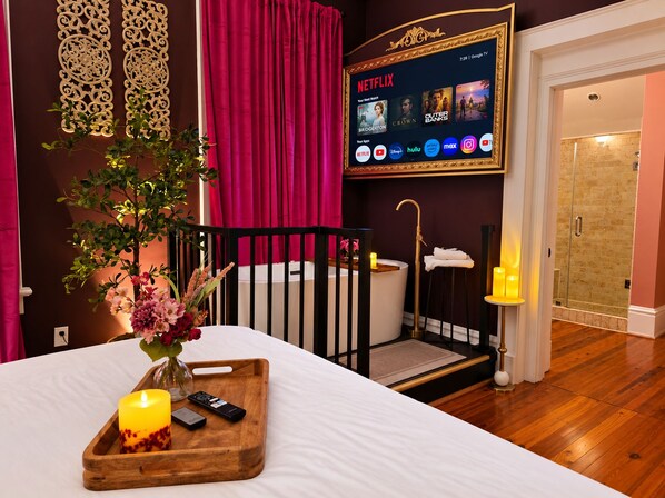 A hotel room with a bed, a TV displaying Netflix, a bathroom with a shower, and a wooden tray with a candle and remote controls.