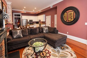 A living room with a sectional sofa, a glass coffee table with flowers, a patterned rug, and a kitchen in the background. - No Car Needed+King Bed+Balcony - Abercorn and York (Savannah)