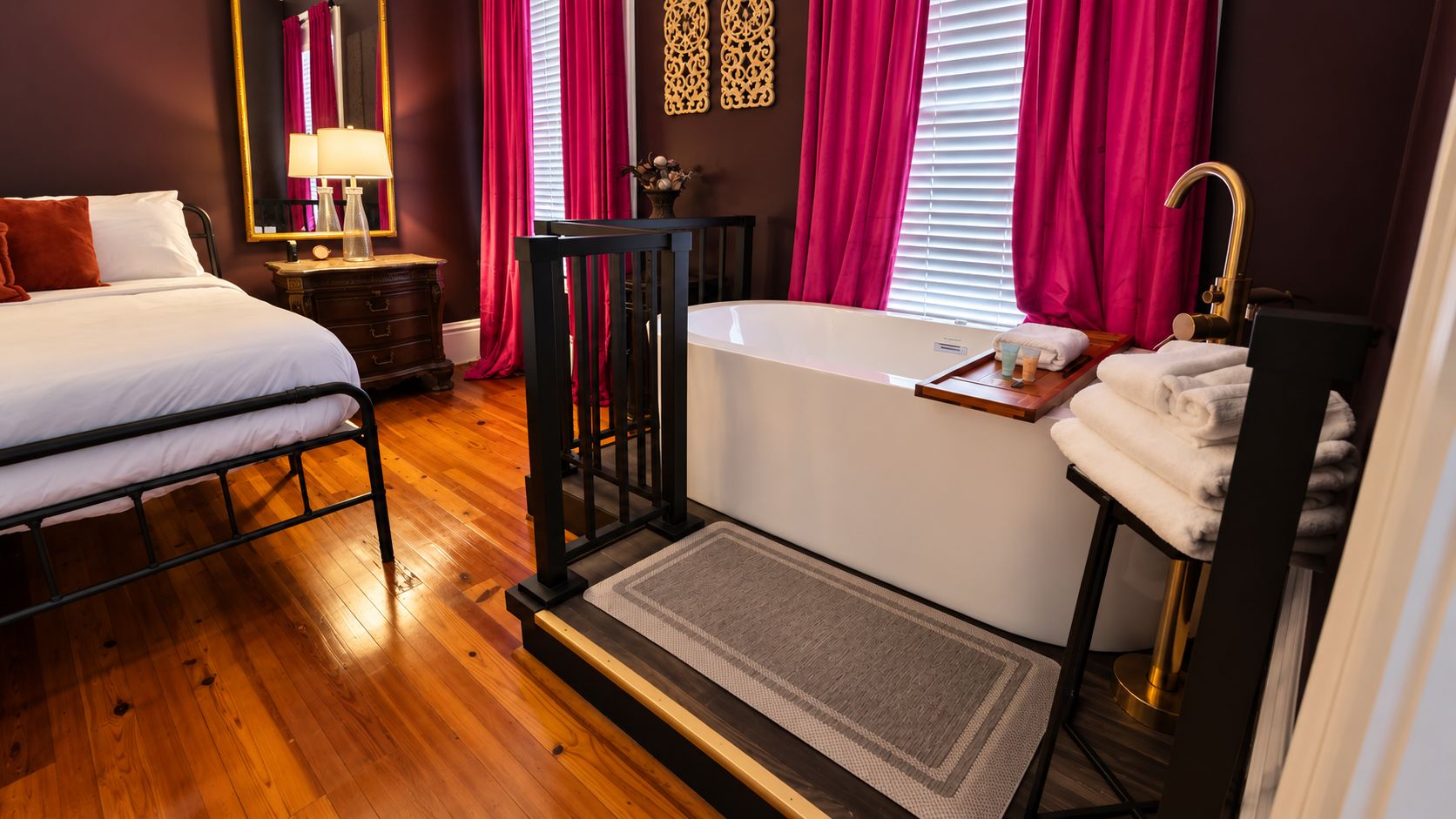 A bedroom with a bed, a bedside table, a lamp, a window with curtains, a bathtub, and a towel rack.