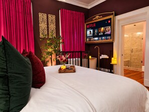 Habitación de hotel con una cama grande, un televisor en la pared mostrando la interfaz de Netflix, una mesita con un jarrón de flores y una zona de ducha visible a través de una puerta abierta.