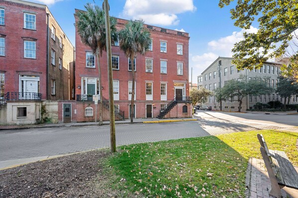 A red brick building with a central staircase, a palm tree, and a grassy area with a bench.