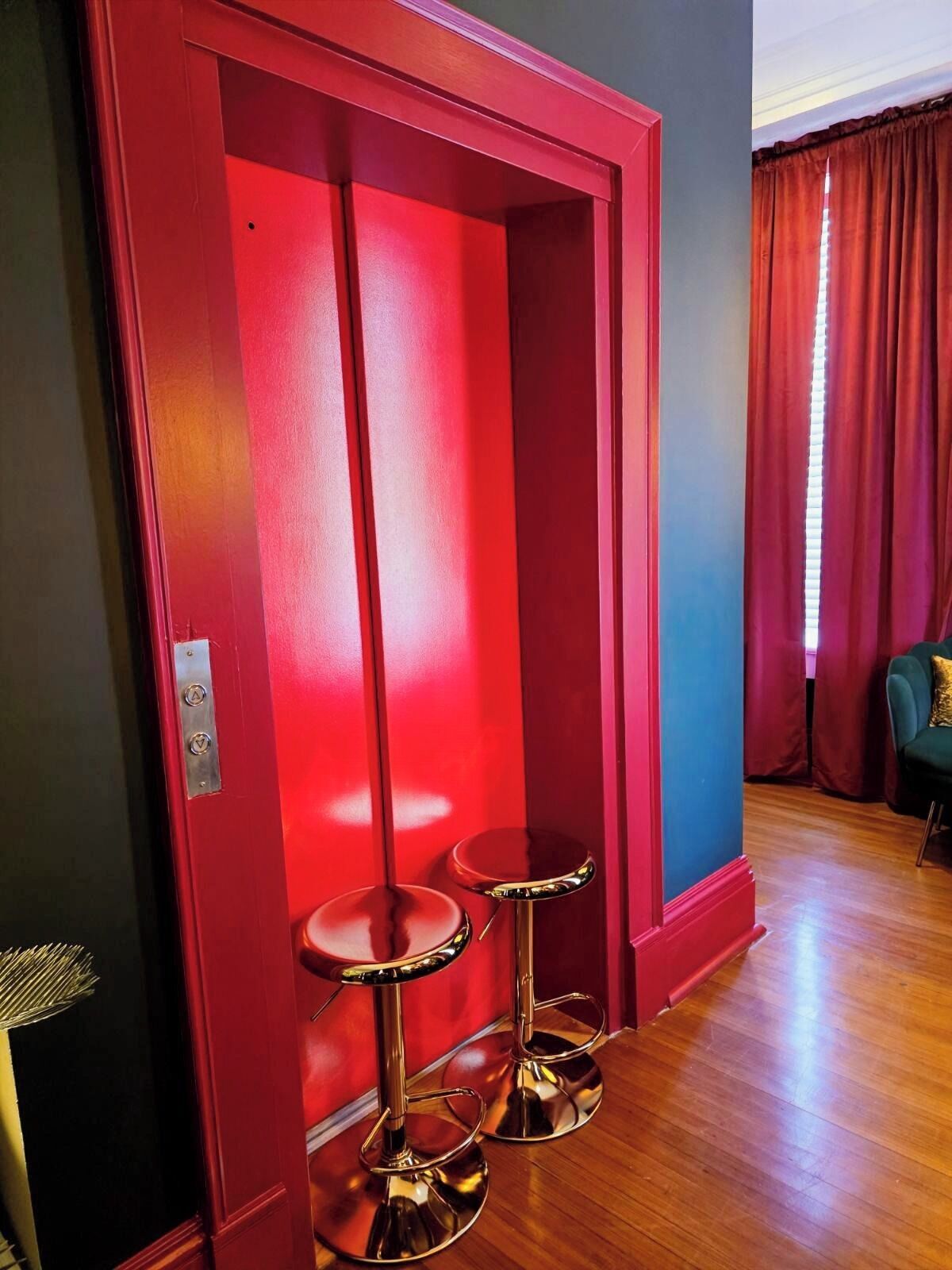 Habitación con un ascensor rojo, dos taburetes dorados, una silla verde y una ventana con cortinas rojas.