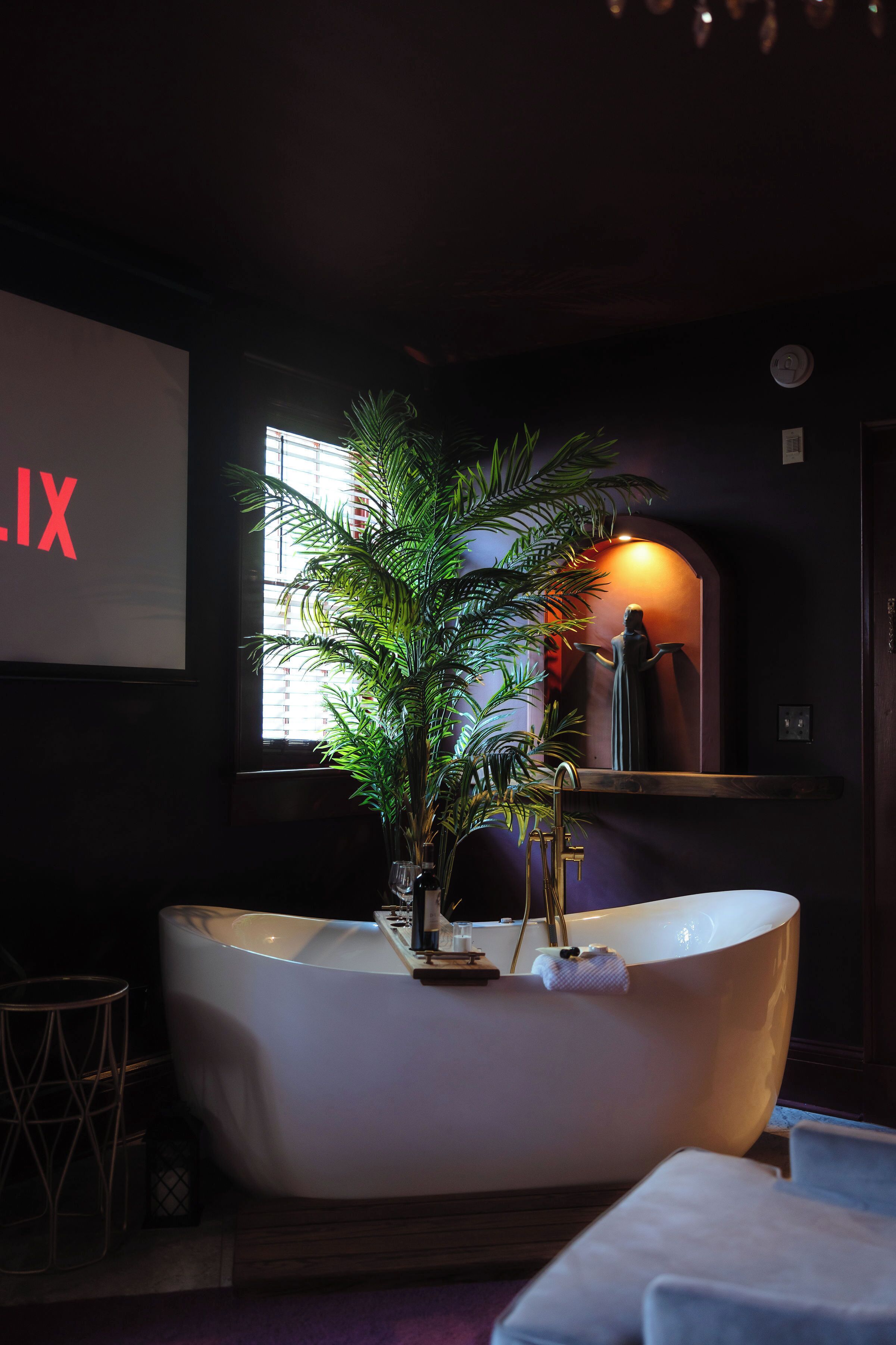 A bathtub with a plant, a sink, and a shelf with a statue in a dimly lit room.