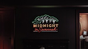 Ein dunkler Raum mit einem Neonzeichen, das 'MIDNIGHT in Savannah' zeigt, über einem Kamin.
