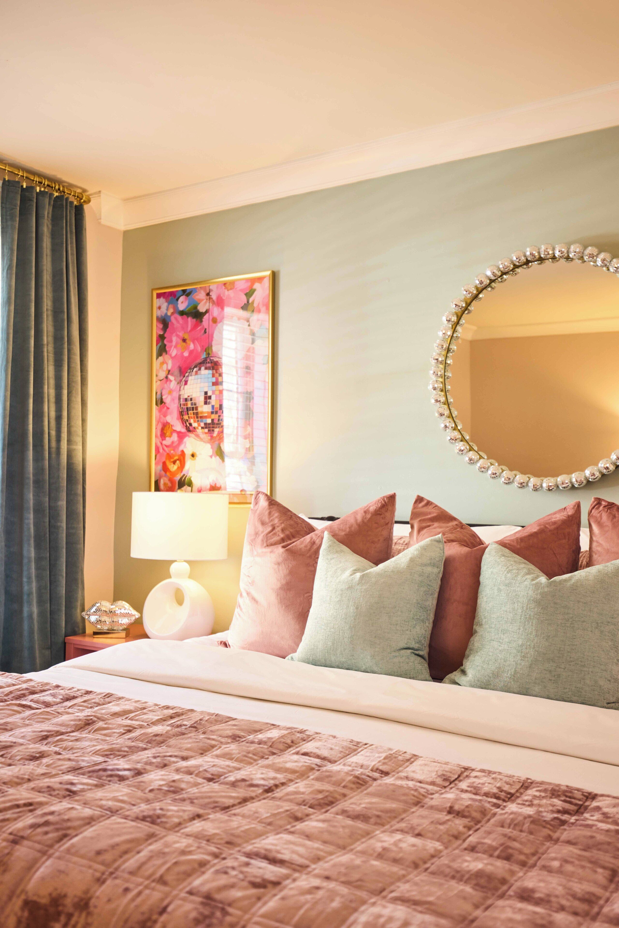 Habitación de hotel con cama, almohadas, lámpara, espejo y un cuadro floral en la pared.