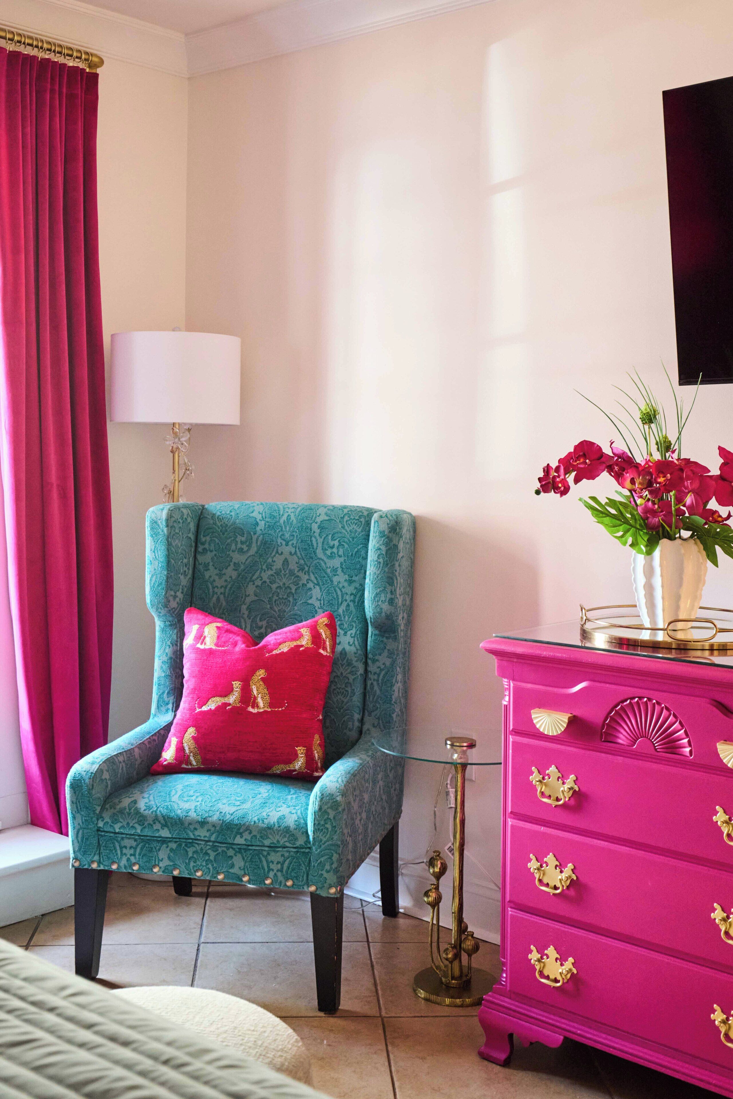 A teal armchair with a red cushion, a pink dresser with gold handles, and a vase of flowers on the dresser.