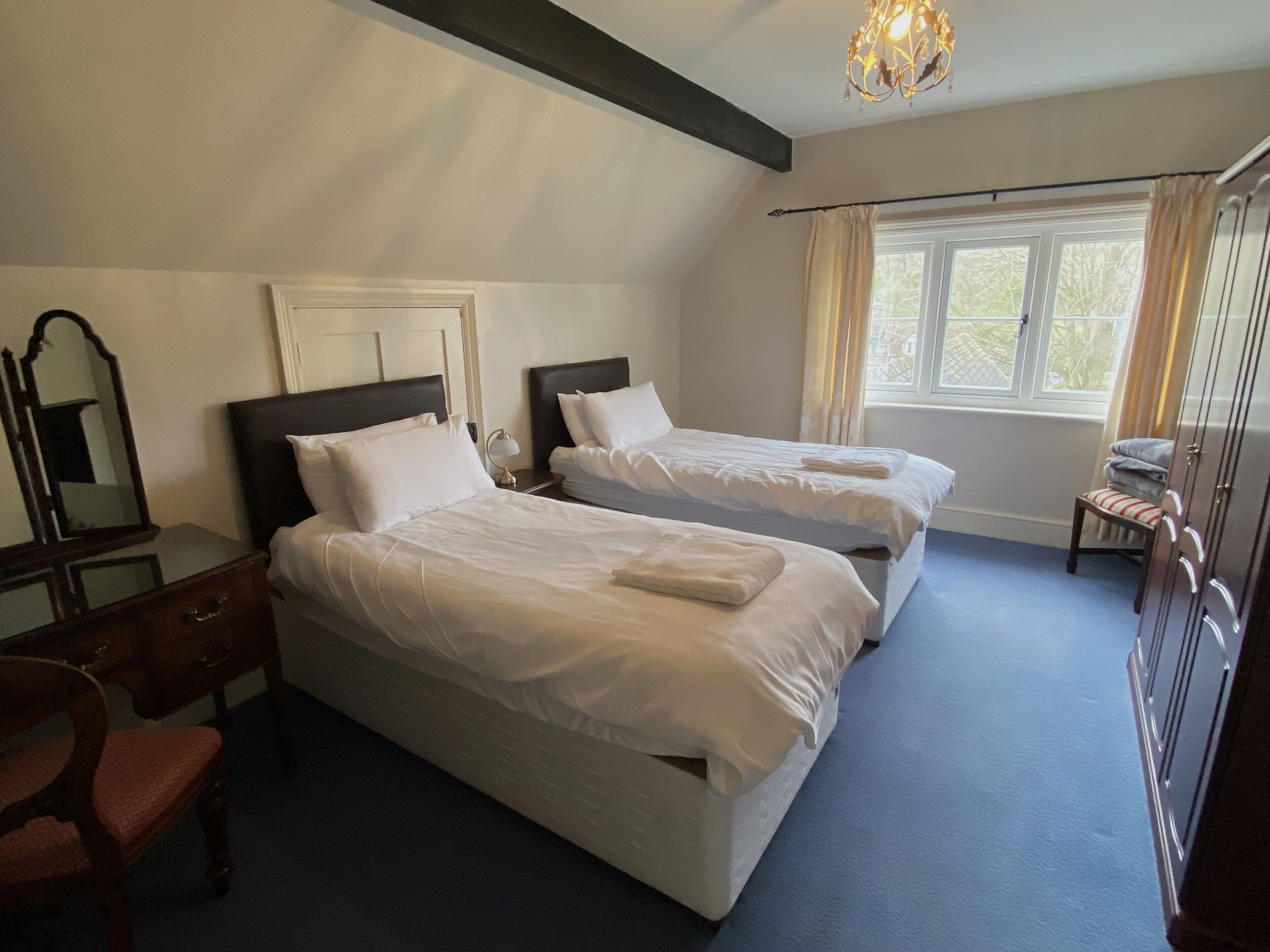 A hotel room with two beds, a dresser, a chair, a window with curtains, and a chandelier.