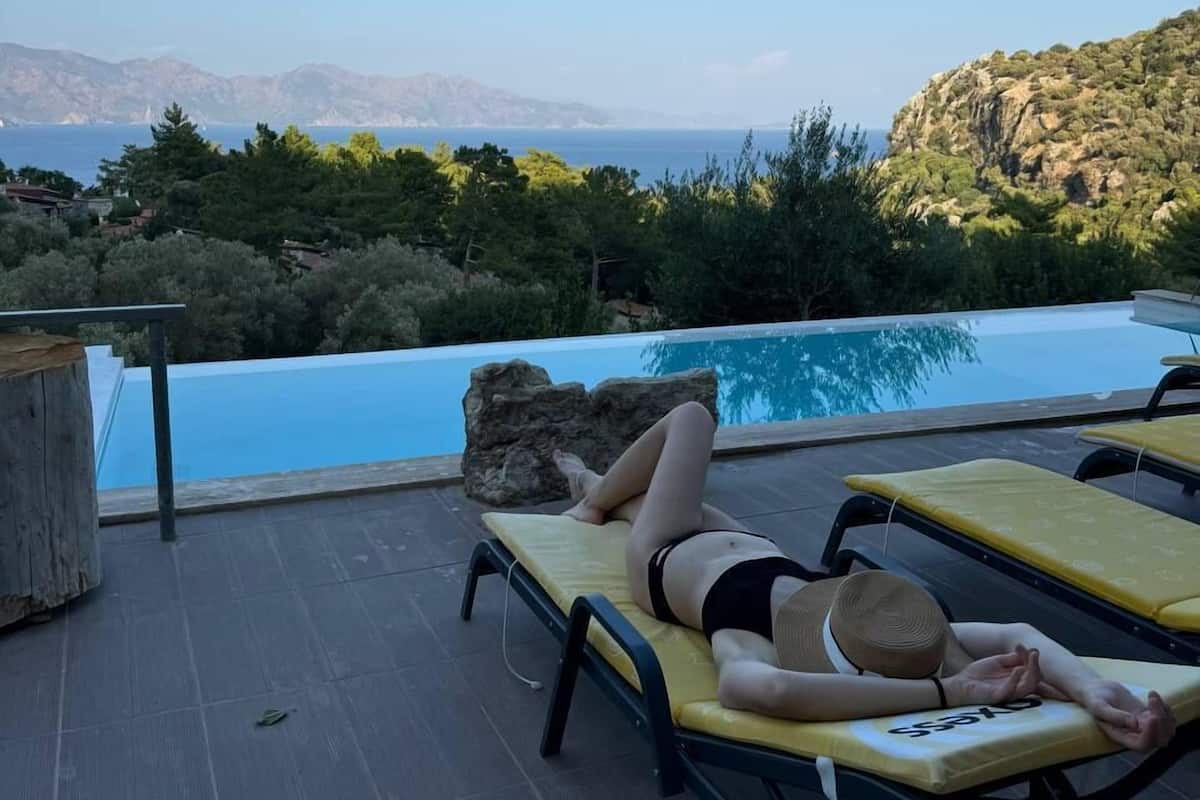 A person relaxing on a sun lounger by a pool with a view of the sea and mountains.