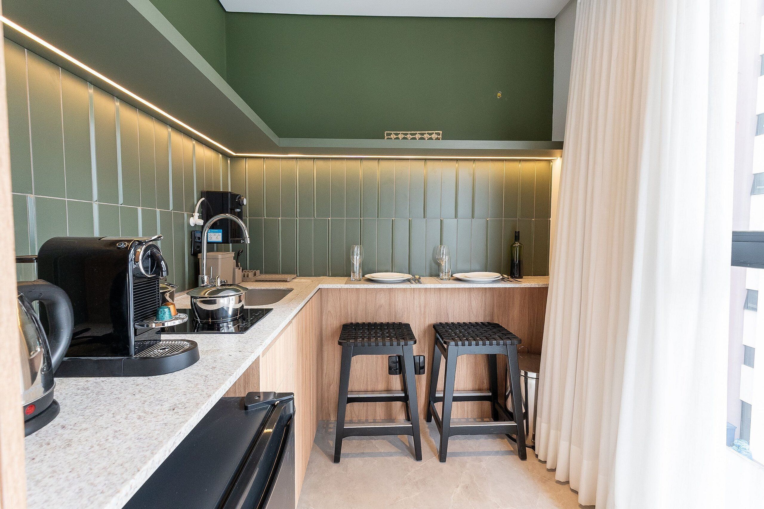A compact kitchen with a coffee machine, a kettle, and a toaster. There are two stools and a sink.