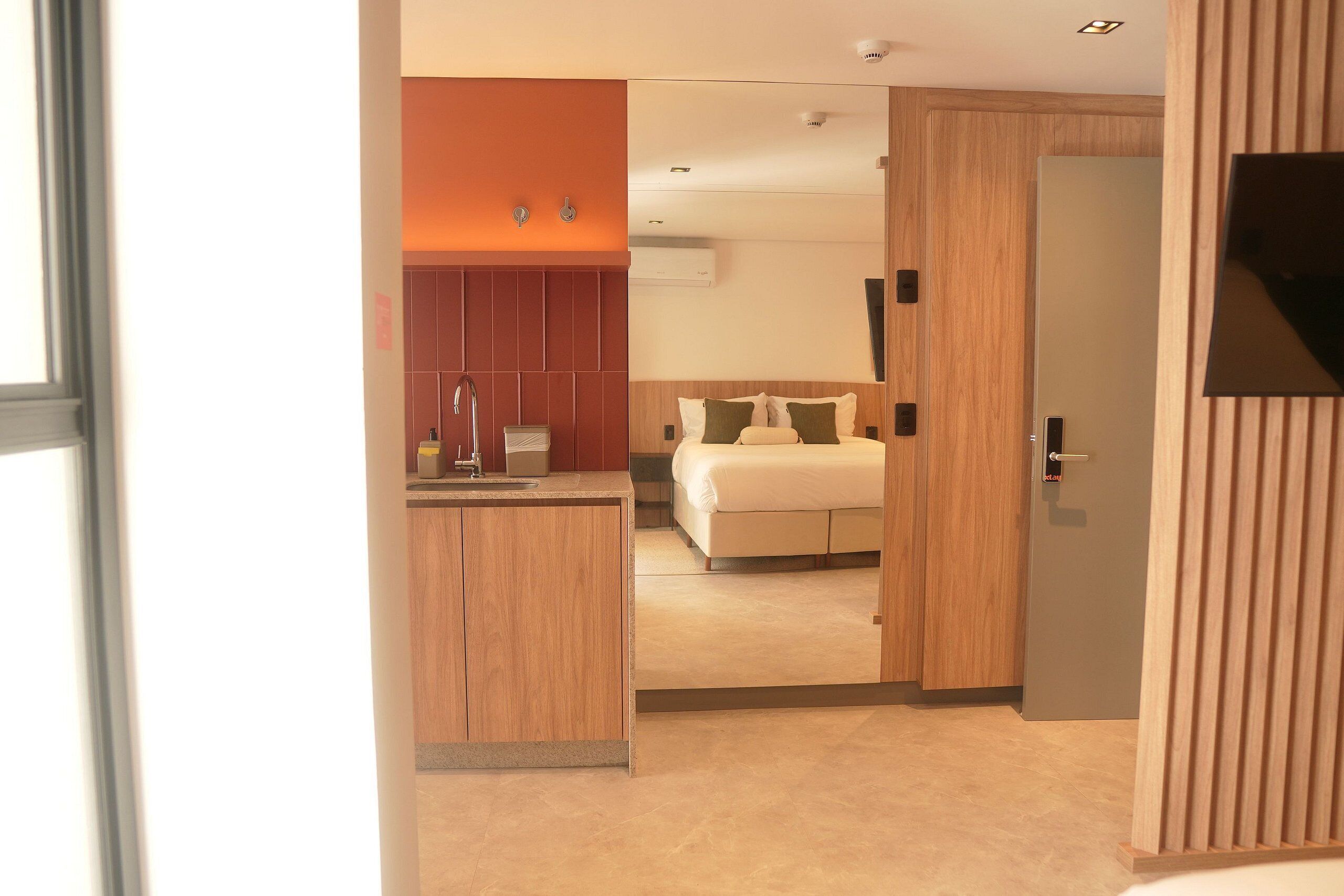A modern hotel room with a wooden vanity, a bed with green and beige bedding, and a wooden door.