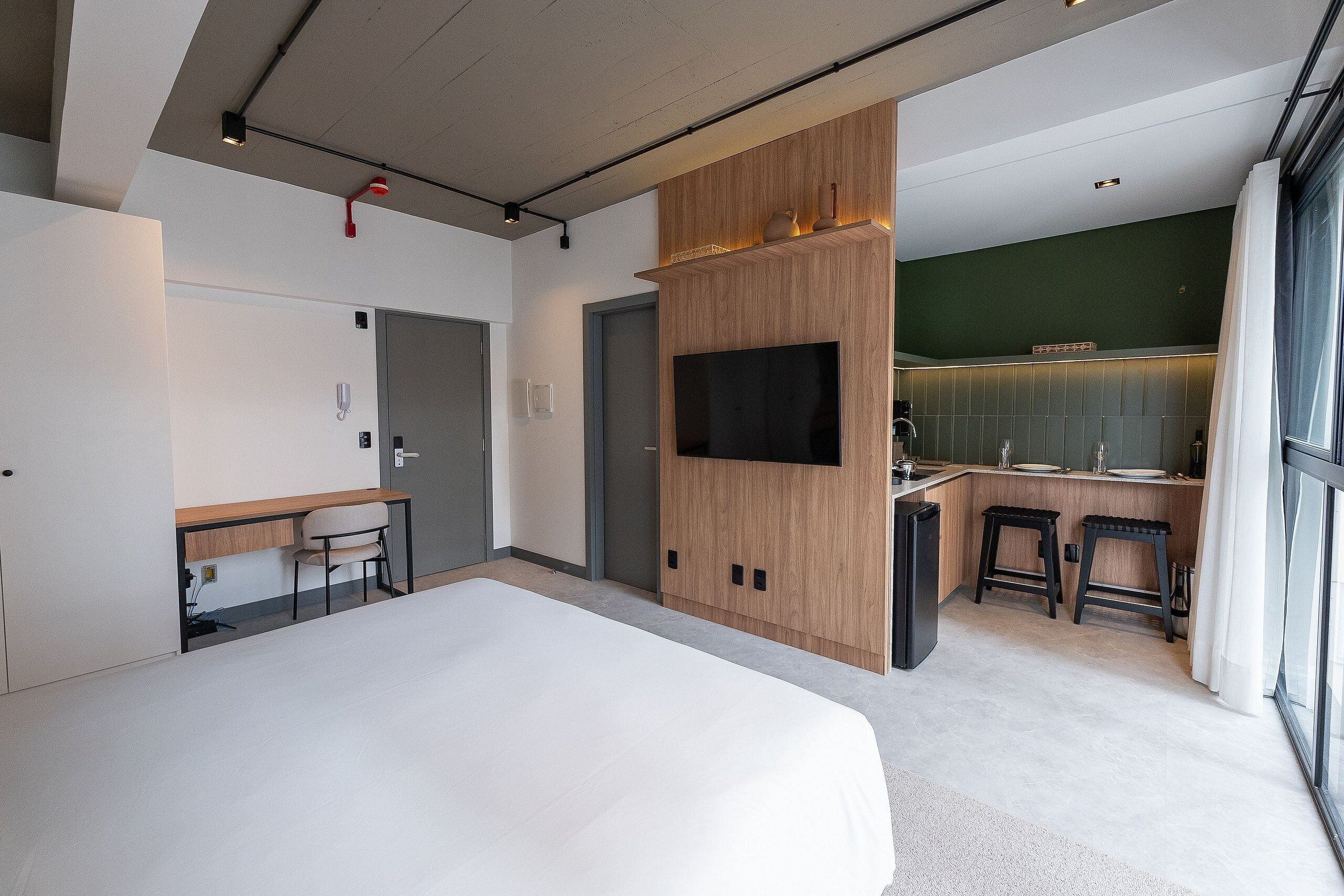 Una habitación de hotel moderna con una cama grande, un escritorio con silla, un televisor montado en un panel de madera y una pequeña kitchenette con taburetes.