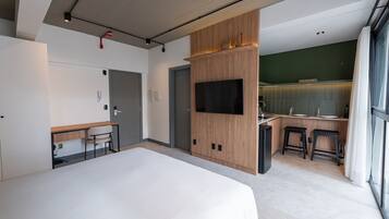 Una habitación de hotel moderna con una cama grande, un escritorio con silla, un televisor montado en un panel de madera y una pequeña kitchenette con taburetes.