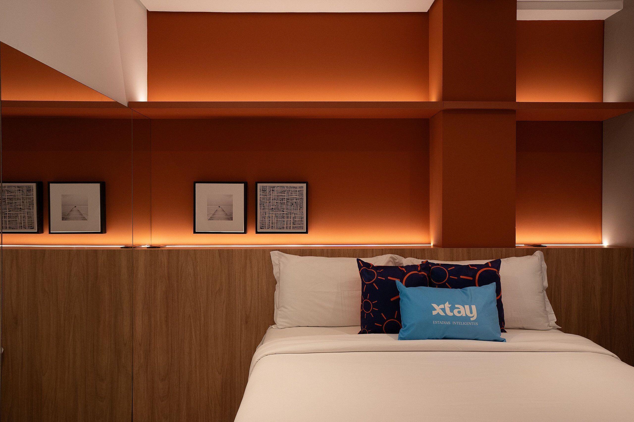 A hotel room with a wooden headboard, a bed with white bedding, and a blue 'xtay' pillow.