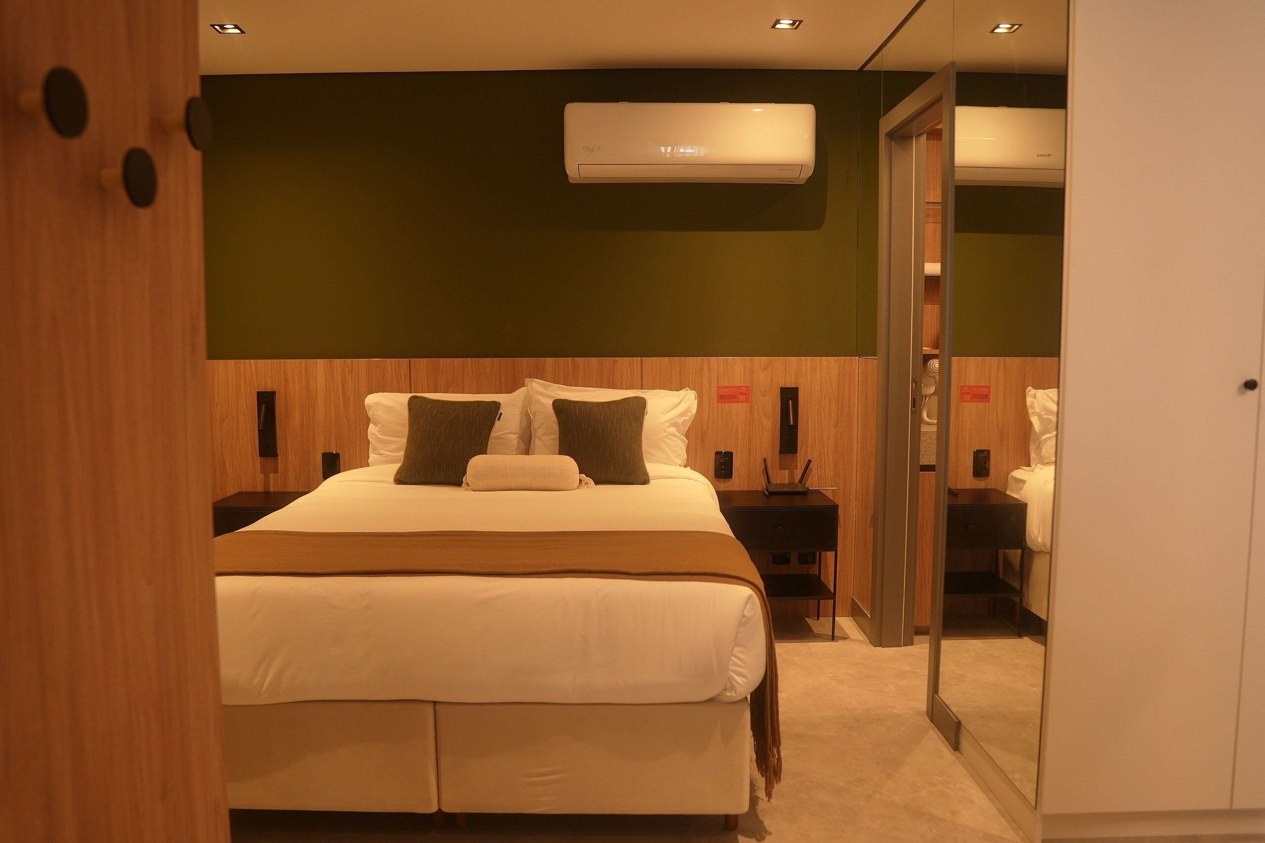 Una habitación de hotel con una cama, mesitas de noche, un equipo de aire acondicionado y un espejo.