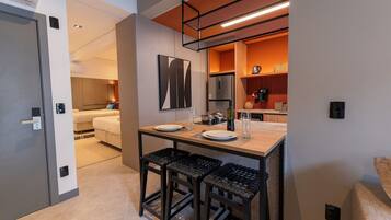 Habitación de hotel compacta con kitchenette, comedor con mesa y sillas, y una cama al fondo.