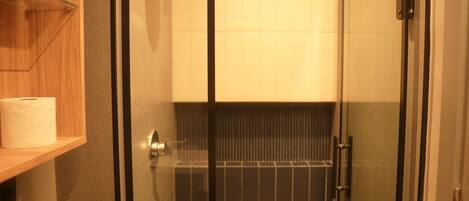 A glass-enclosed shower with a tiled wall, a wooden shelf with a roll of toilet paper, and a door with a handle.