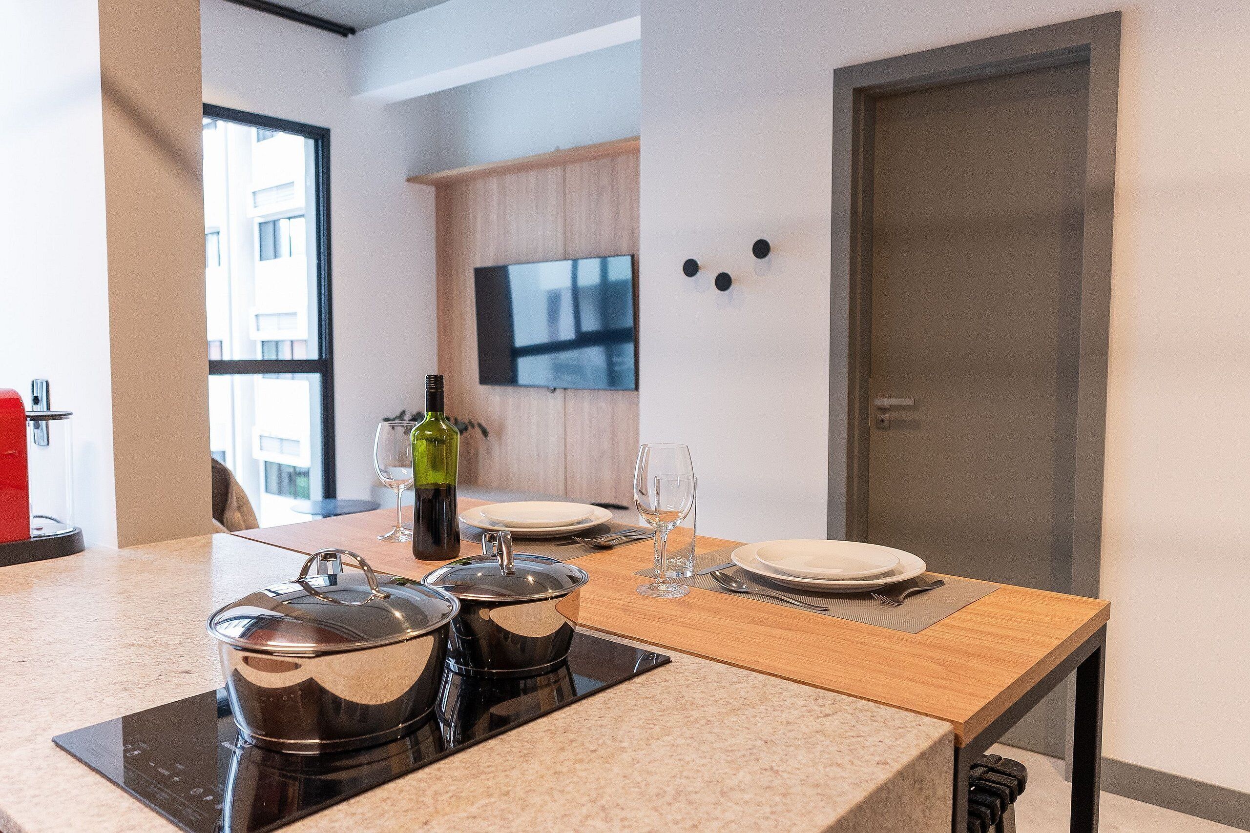 Una cocina moderna con una mesa de madera preparada para dos comensales, un televisor montado en la pared y una puerta a la derecha.