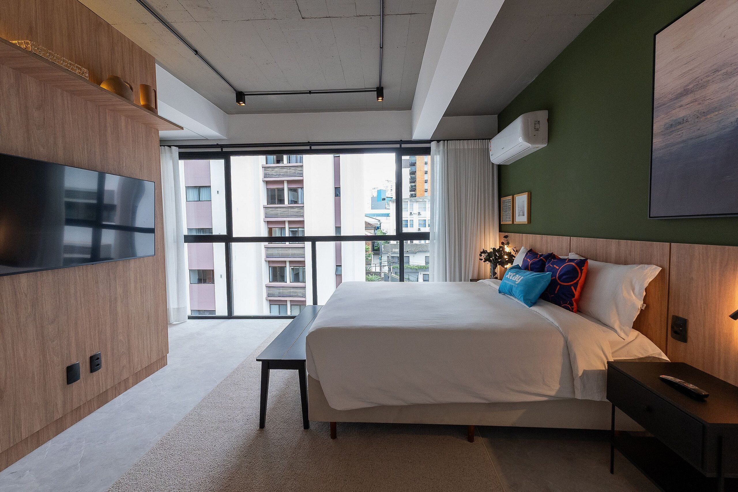 Een moderne hotelkamer met een groot bed, een flatscreen televisie, een nachtkastje en uitzicht op de stad door grote ramen.