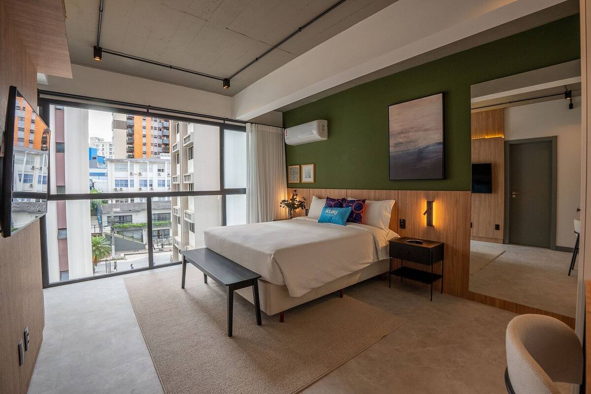 Ett modernt hotellrum med en stor säng, ett skrivbord och utsikt över staden genom stora fönster.