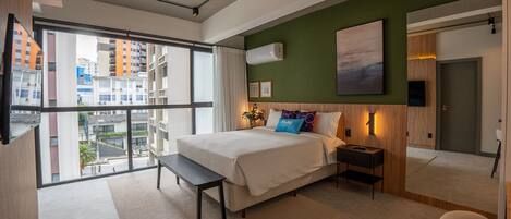 A modern hotel room with a large bed, a desk, and a view of the city through large windows.