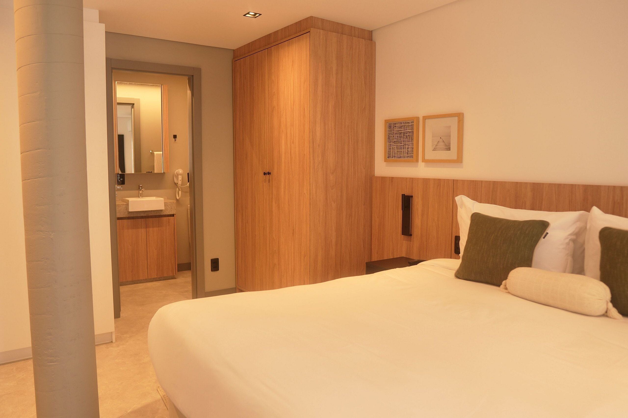 A hotel room with a large bed, a wooden wardrobe, a bathroom with a sink, and two framed pictures on the wall.