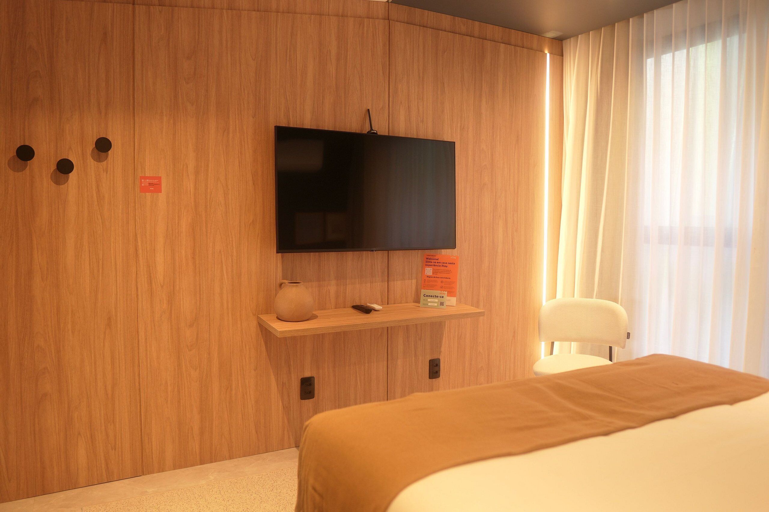 Habitación de hotel con una cama, un televisor montado en una pared de paneles de madera, un estante pequeño con un jarrón y un control remoto, y una silla junto a una ventana con cortinas.