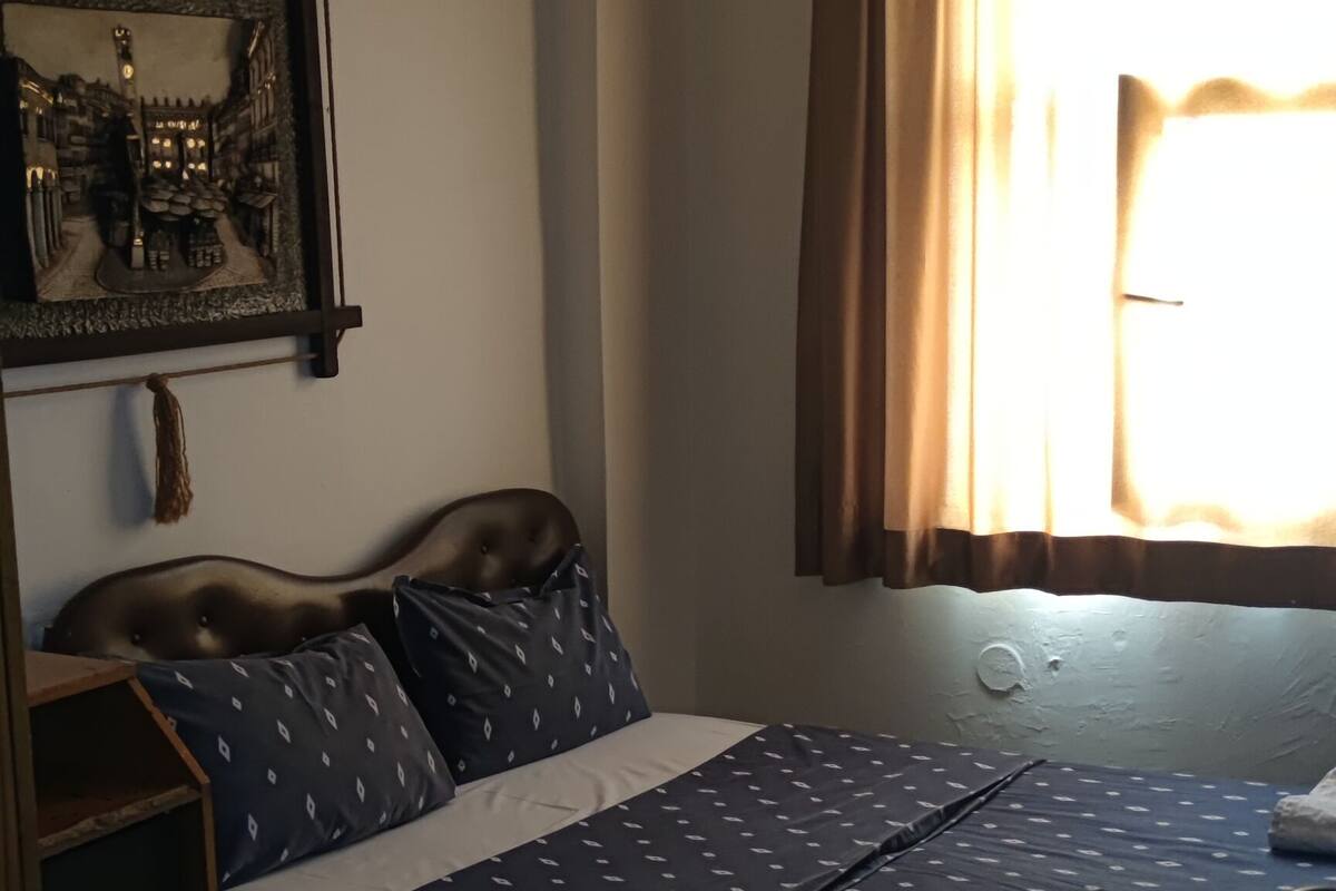 A bedroom with a bed, a nightstand, a wall-mounted shelf, a window with curtains, and a patterned rug.