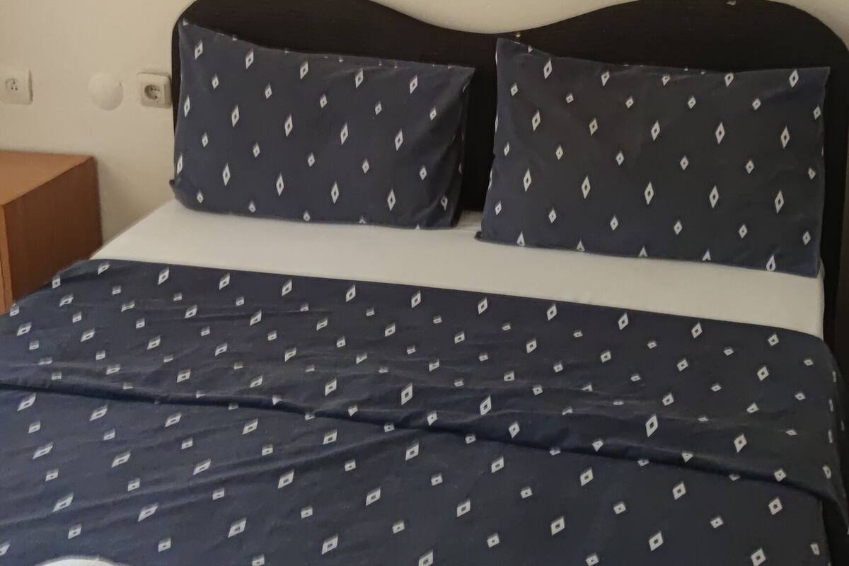 A bed with a patterned bedspread and pillows, a wall-mounted painting, and a small wooden nightstand.