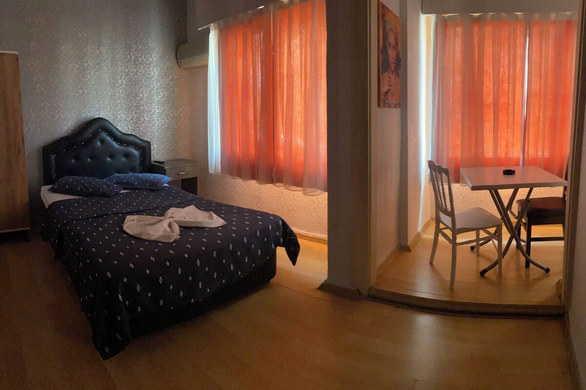 A bedroom with a bed, a wooden wardrobe, a small table with a chair, and a window with orange curtains.