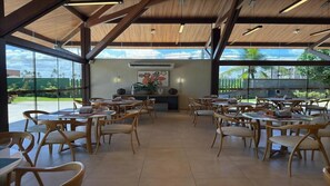 A spacious indoor dining area with wooden tables and chairs, large windows offering a view of palm trees and a clear sky. - Malawi Beach Resort (Ipojuca)