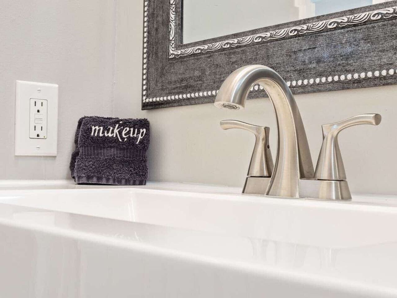 A bathroom with a white sink, a silver faucet, a black towel with 'makeup' written on it, and a mirror with a decorative frame.