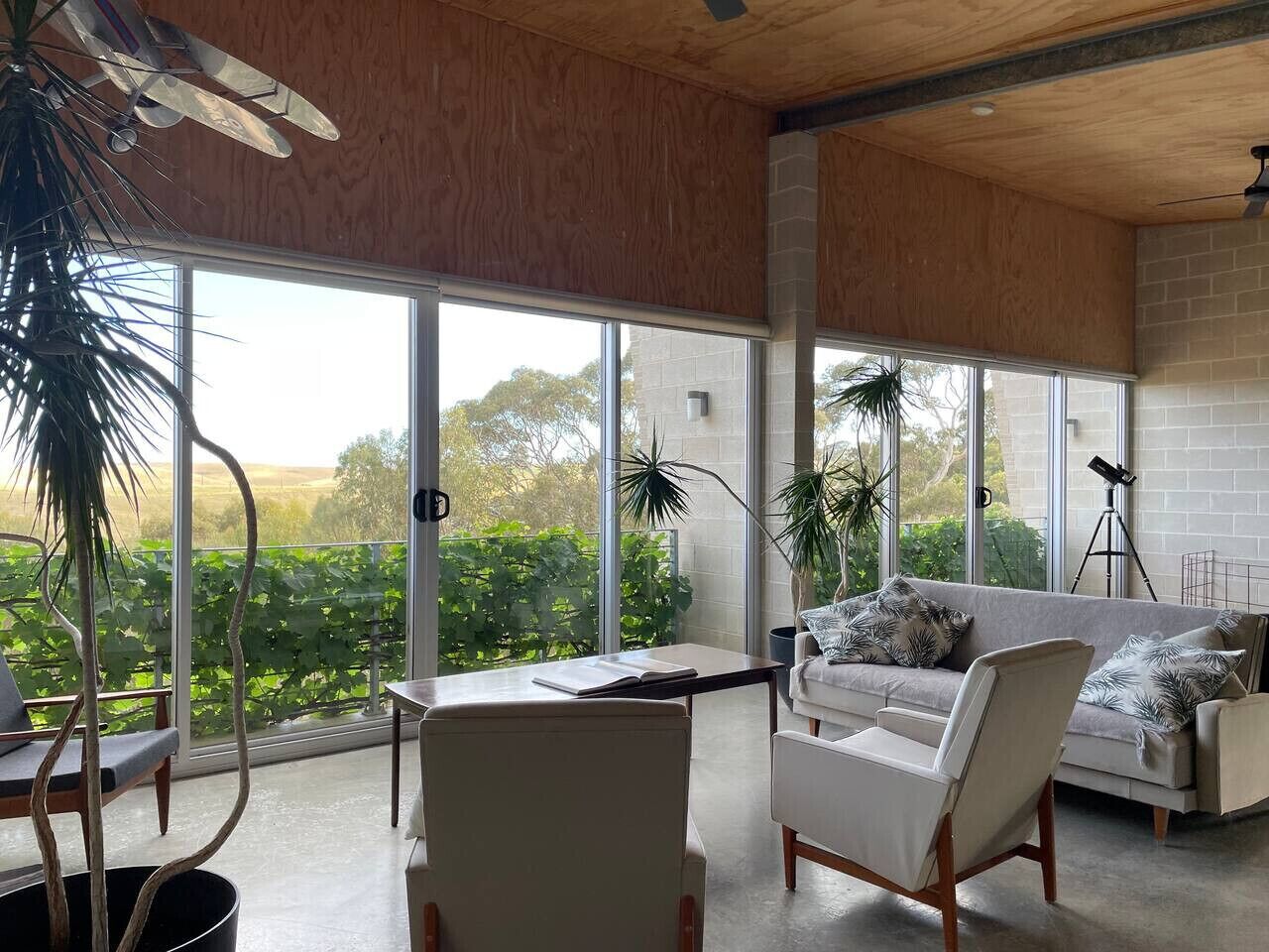 A stylish, modern living room with a wooden ceiling and large sliding glass doors that offer a view of greenery outside. The room is furnished with comfortable seating including a sofa with patterned cushions and several armchairs, a coffee table, and is 