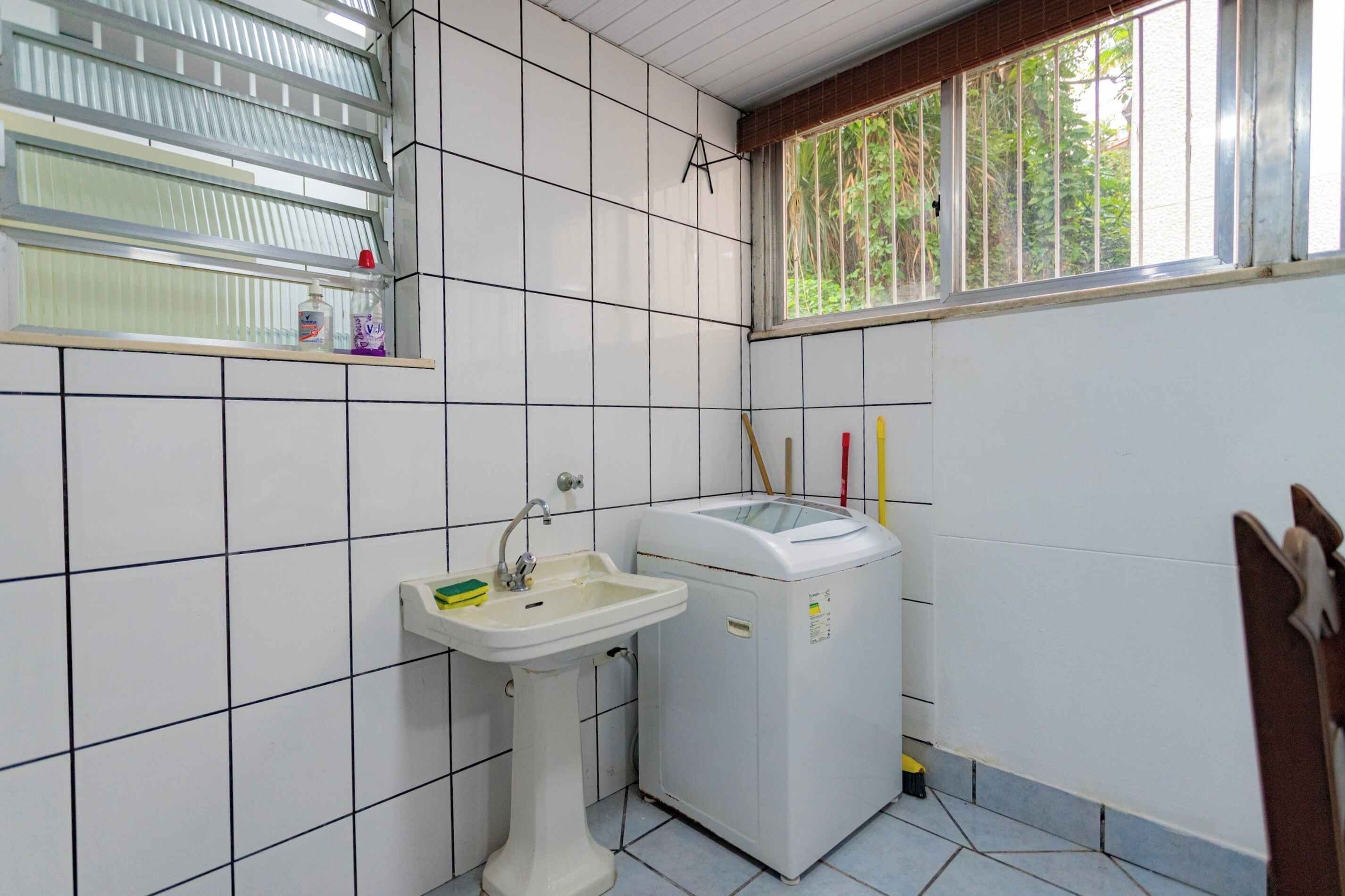 A small bathroom with a sink, a washing machine, a window with a view of greenery, and a shelf with cleaning supplies.