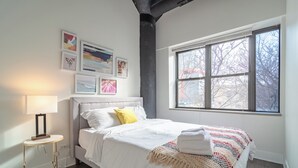 Een slaapkamer met een bed, een nachtkastje met een lamp, een raam met uitzicht en wanddecoratie.
