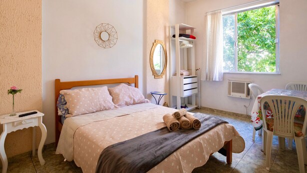 Un dormitorio con cama, mesita de noche, espejo, armario, ventana y una mesa con sillas.