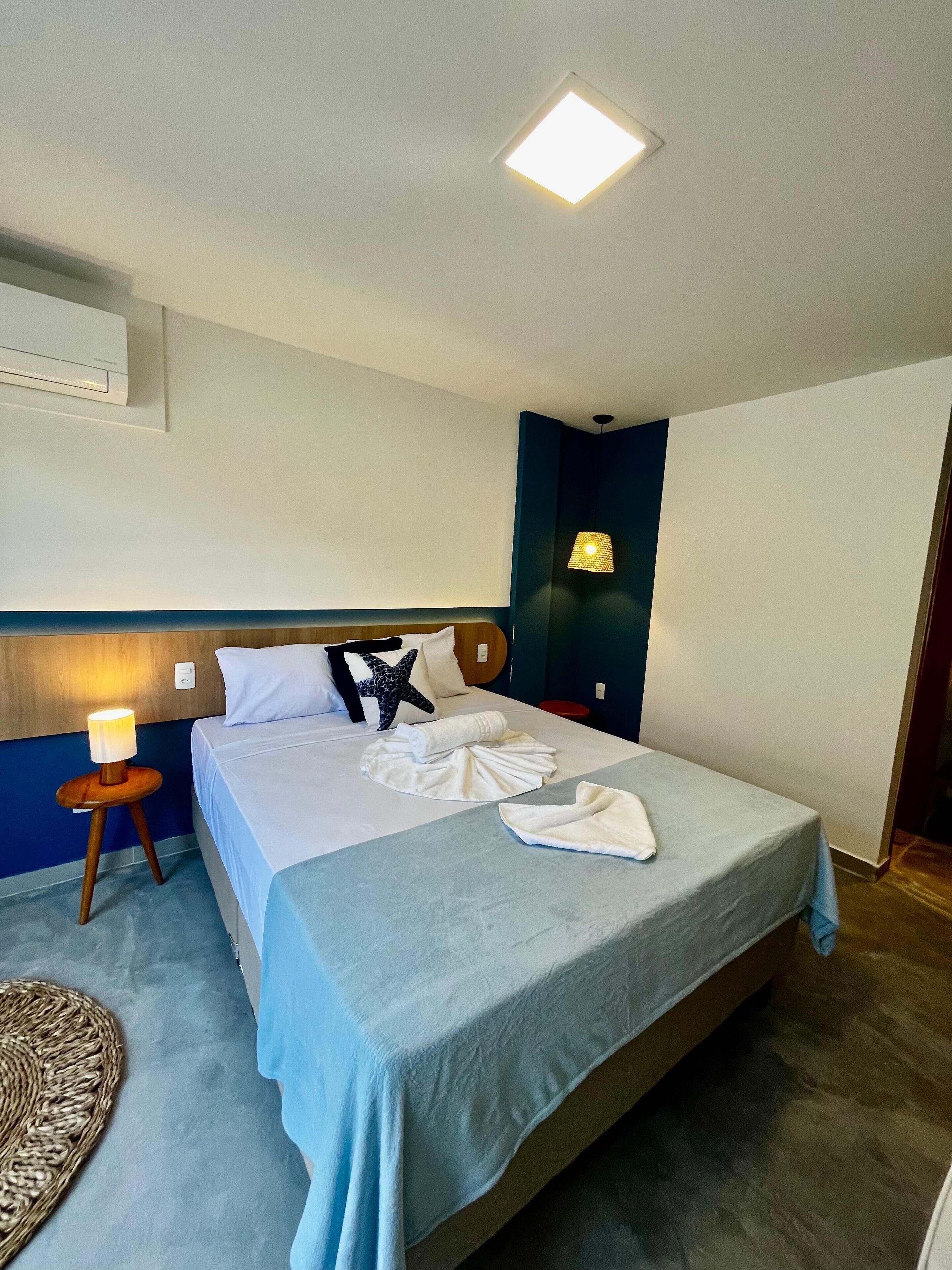 Une chambre d’hôtel comprenant un lit, une lampe de chevet, un climatiseur fixé au mur et une petite table avec une lampe.