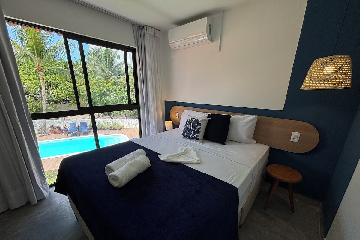 A hotel room with a bed, a small stool, a window with a view of a pool, and an air conditioning unit on the wall.