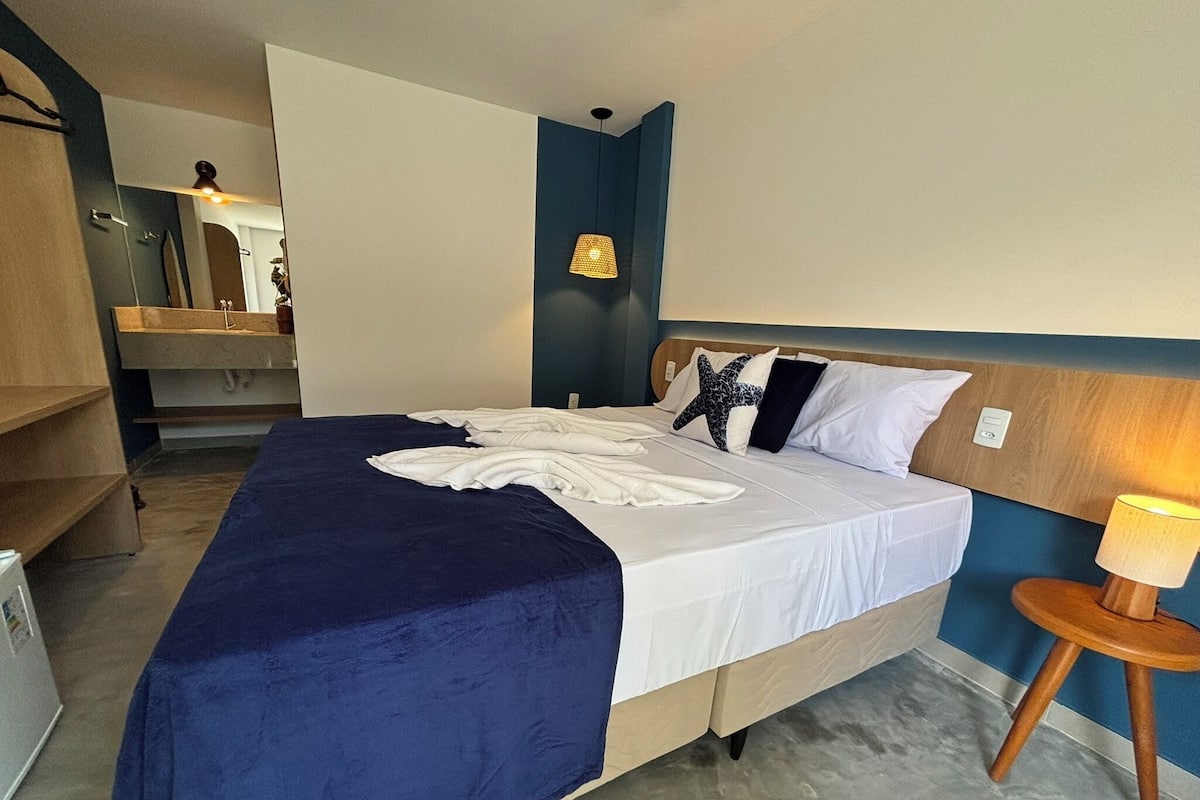 A hotel room with a large bed, a bedside table with a lamp, a mirror, and a small seating area with a chair.