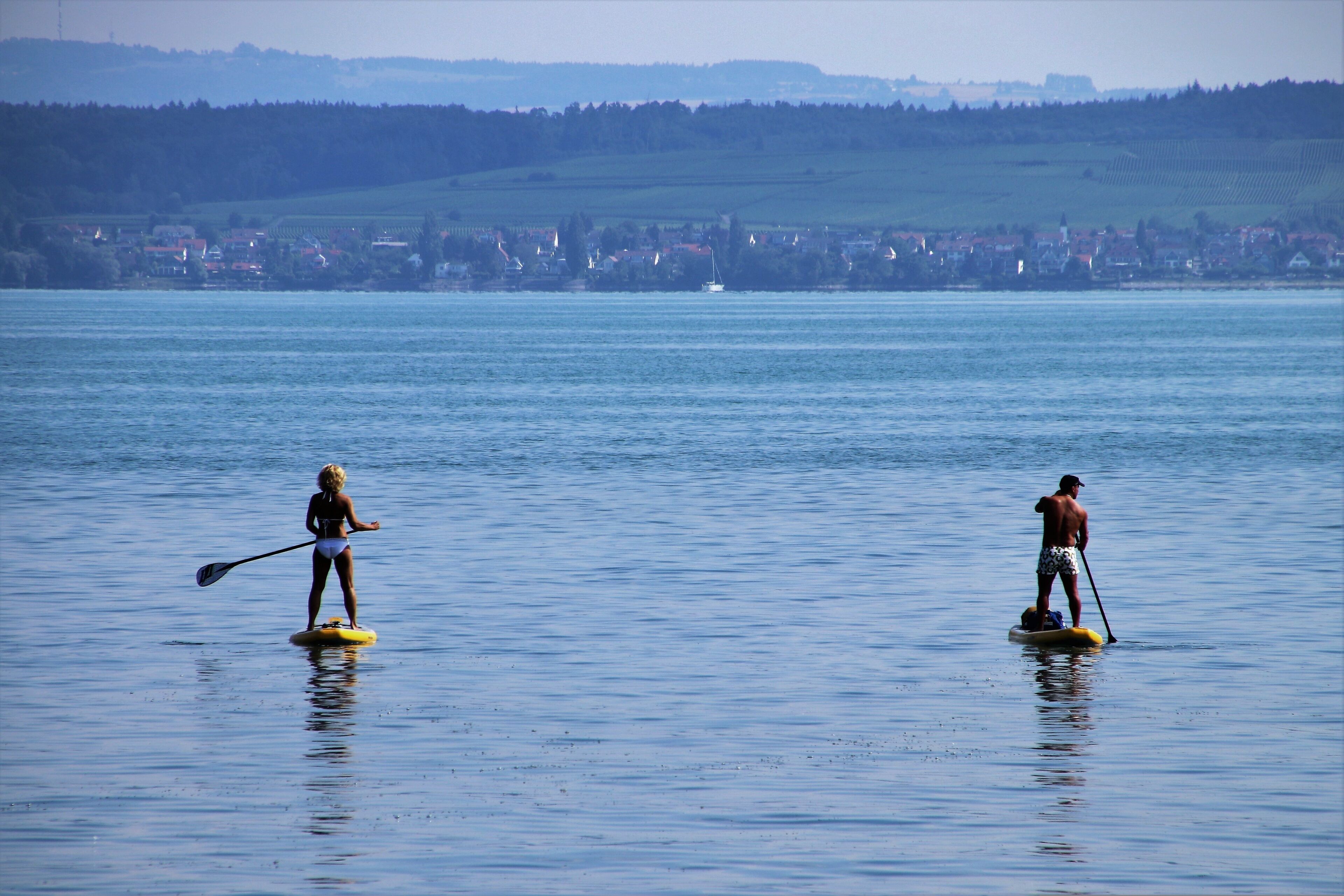 Zwei Personen paddeln auf einem ruhigen See, im Hintergrund eine Stadt.